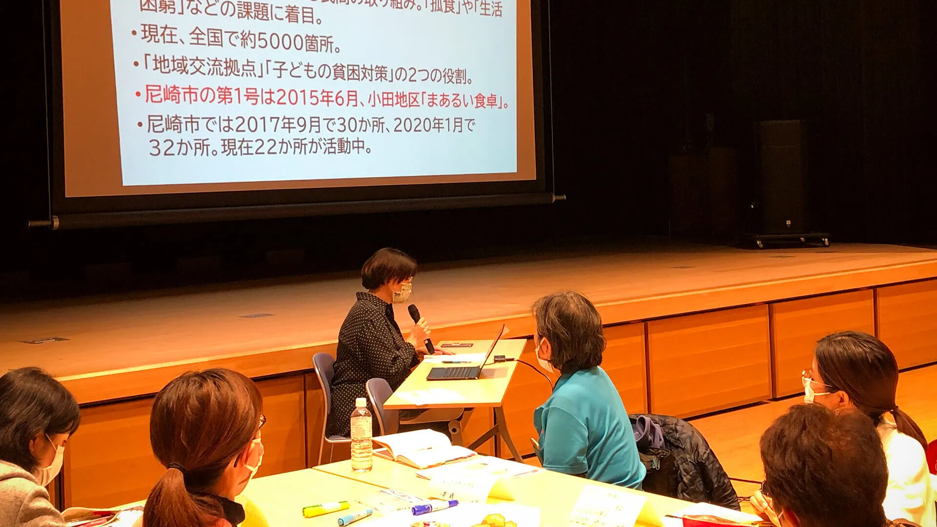 写真：スクリーンに写したスライドを見ながら、マイクを持った方の声に耳を傾ける椅子に座った方達